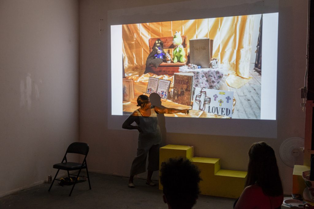 Image: A photo of the second session of CANJE. Rainn Thomas stands in the front of a room while pointing at their work, which is being projected onto the wall behind her. Photo by Ryan Edmund Thiel.