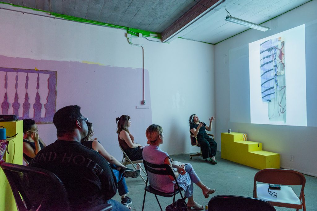 Image: A photo of the first session of CANJE. The presenting artist Victoria Martinez sits in a chair in the front of the room while gesturing towards her presentation on the wall. She is talking about her work with the CANJE cohort and special guest, Francine Almeda. Photo by Ryan Edmund Thiel.