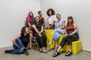 Image: A photo of the first session of CANJE. The following folks sit and stand together while smiling at the camera: Victoria Martinez, Morgan Green, Francine Almeda, Natalia Villanueva Linares, Kiki Lechuga-Dupont, EdVette Jones, Tempestt Hazel, and Christina Nafziger. Photo by Ryan Edmund Thiel.