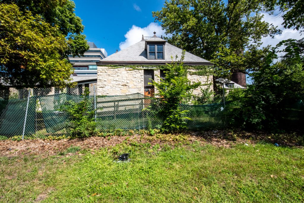 Image: A once-abandoned structure purchased by the DuSable Museum is surrounded by a green fence and trees, with a modern structure in the background. Photo by EdVetté Wilson Jones.