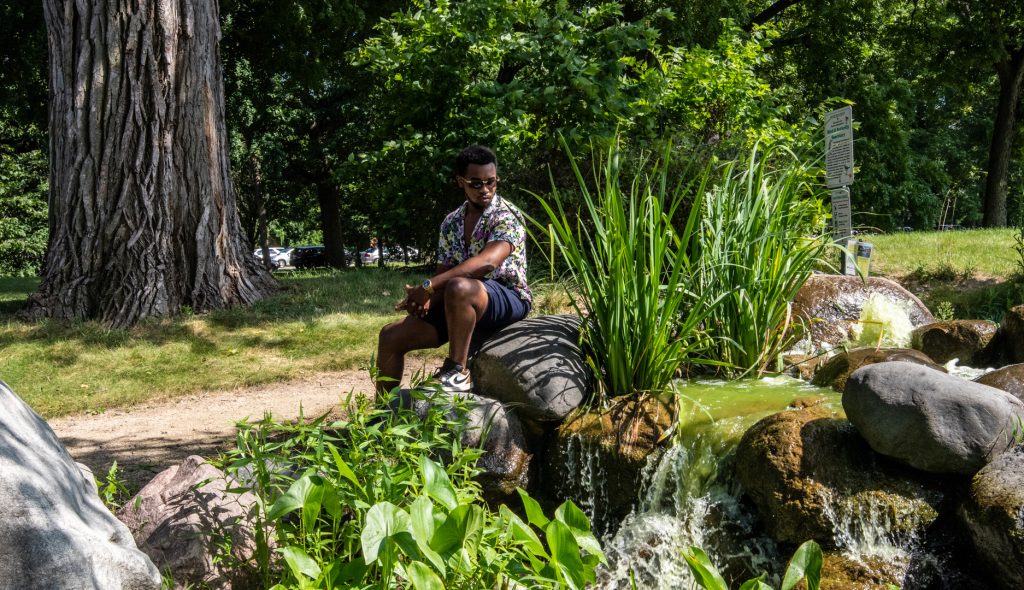 Image: A park-goer relaxes beside the man-made waterfall built by the sons of Frederick Law Olmsted. The waterfall is surrounded by large rocks and various types of lush green foliage. Photo by EdVetté Wilson Jones.