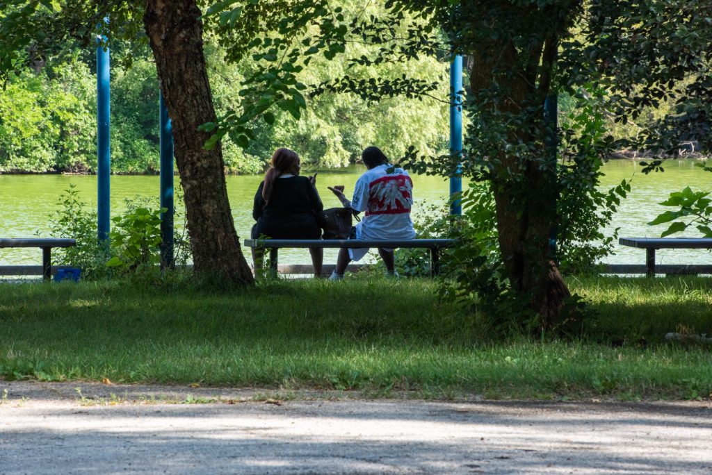 Image: Two lovers sit and talk at Bynum Island in the park. They are on a bench overlooking water and lush green foliage. Photo by EdVetté Wilson Jones.