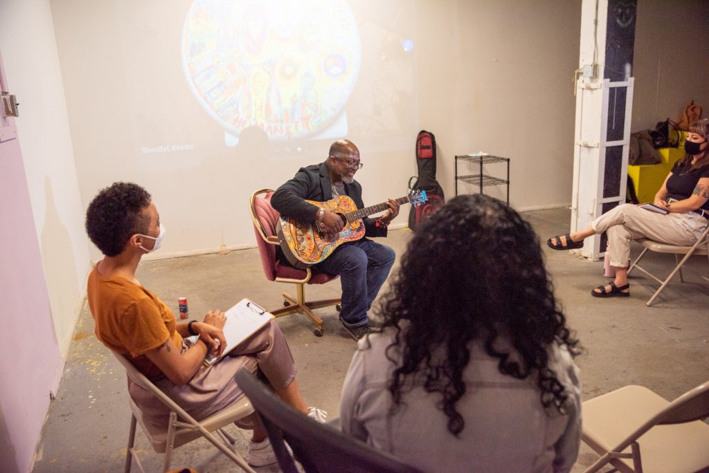 Image: A photo of the fourth session of CANJE. Presenting artist Carnell Newbill (aka: Spike Rebel) sits in a chair in the middle of the room while singing and play guitar. Around him sits members of the CANJE cohort. The special guest was Yvonne Toney. Photo by EdVette Jones.