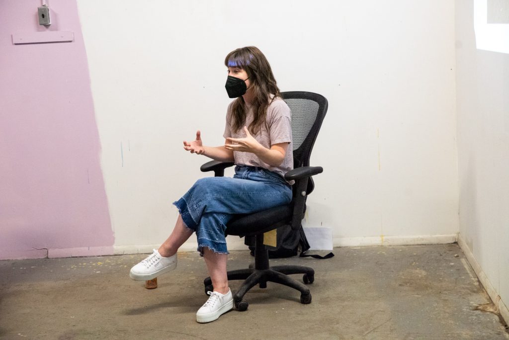 Image: A photo of the fifth session of CANJE. Presenting artist Kelly Kristin Jones sits in a chair in the corner of the room while gesturing and talking about her artwork. Photo by EdVette Jones.