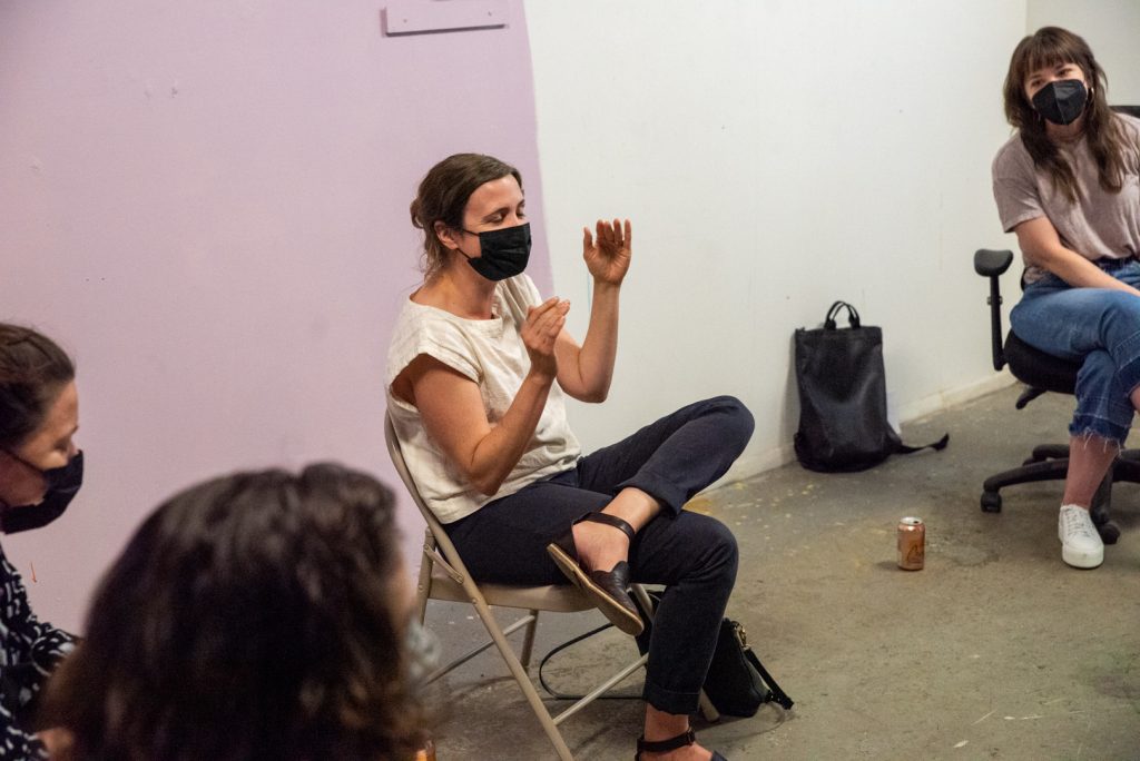 Image: A photo of the fifth session of CANJE. Kristin Taylor sits in a chair while gesturing and talking about the work of Kelly Kristin Jones, who sits to her right. Photo by EdVette Jones.