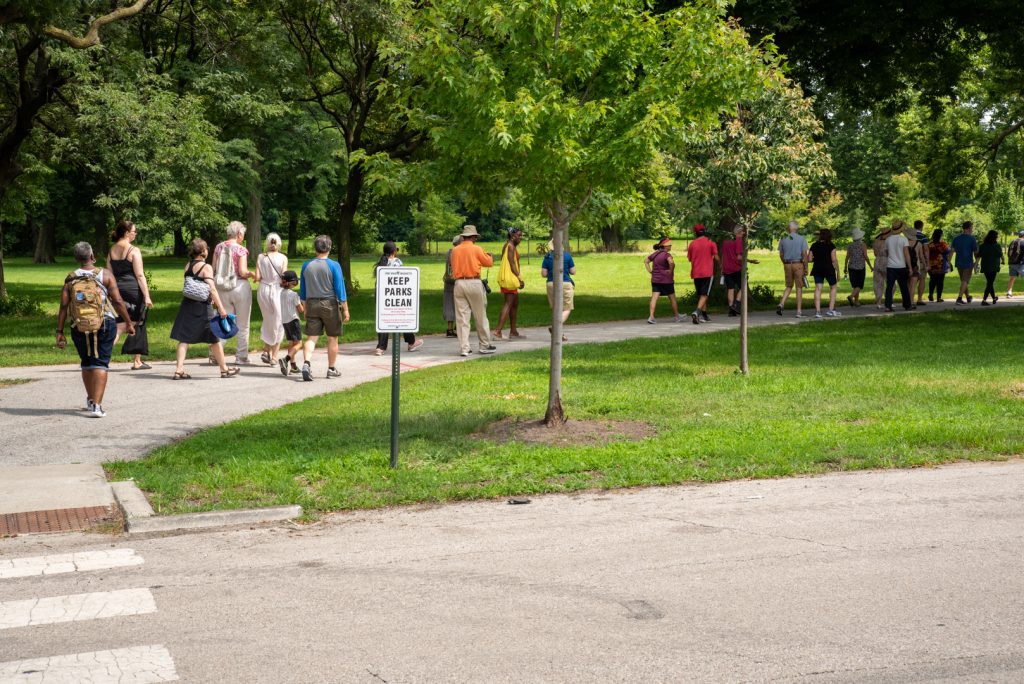 Image: The tour starts as tourists walk along a path with a sign that reads "Keep Parks Clean". The path is partly shaded and surrounded by lush green grass and trees. Photo by EdVetté Wilson Jones.