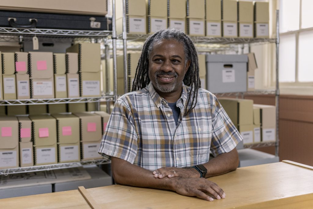 Left Image: Archivist Dino Robinson smiles towards the camera while in the archives of Shorefront Legacy Center. Photo by Ryan Edmund Thiel.
