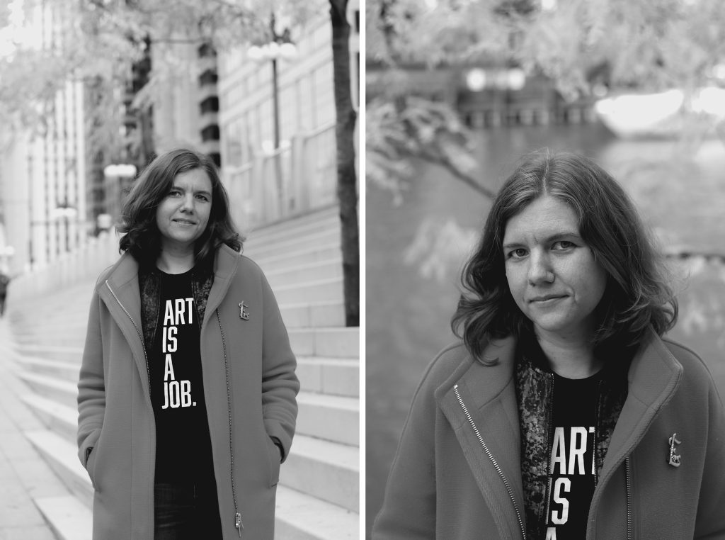 Image: Two black-and-white portraits of Elsa Hiltner, associate director of programs at Lawyers for the Creative Arts and a pay equity advocate who co-founded On Our Team, an organization which works to build pay and labor equity in the theater industry. In both images she wears a heavy overcoat and a t-shirt which says "art is a job". In the left image, she stands along the Chicago Riverwalk on LaSalle Street, while the right image is a more closely-cropped portrait with the river in the background. Photos by zakkiyyah najeebah dumas o’neal.