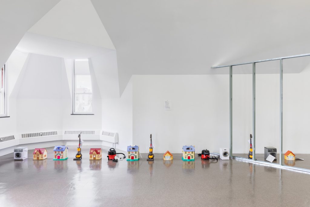 Image: Ghislaine Leung, Public Sculpture, 2018. 16 mini houses and alternating vacuum cleaners lined up in a row. The last three are separated from the rest by a metal frame across the gallery space. Courtesy of the Renaissance Society at the University of Chicago. Photograph by Useful Art Services.