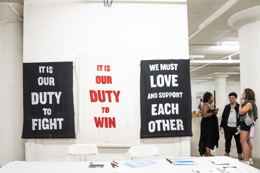 Image: Three banners hung over a table. The banners read "IT IS OUR DUTY TO FIGHT", "IT IS OUR DUTY TO WIN", and "WE MUST LOVE AND SUPPORT EACH OTHER." Photo courtesy of Colectivo Multipolar.