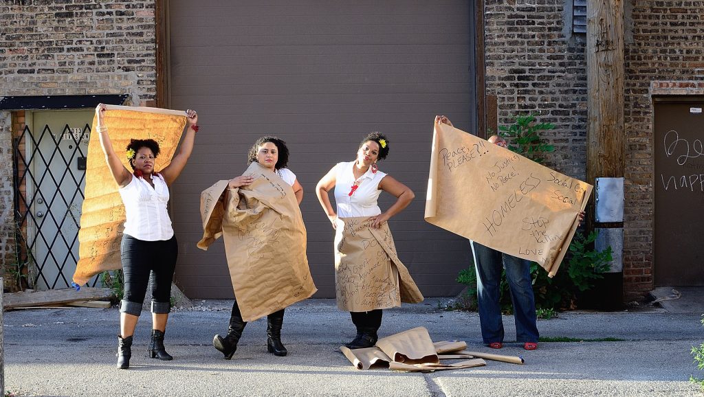 Image: Performers (L-R) Felicia Holman, Meida Teresa McNeal, Aisha Jean-Baptiste, and Abra Johnson, from Honey Pot Performance's project Price Point, June 2013. The project is a multi-arts performance exploring notions of fairness and balance, or the lack thereof, in today’s economic landscape through a mixture of the tragic and comic. The performers are seen in front of a loading dock, holding up brown paper banners with hand-written messages such as "no justice, no peace!", "stop the killing", "be the change", as well as references to public housing, the social contract, imperialism, colonialism, and other topics. More banners are stacked on the ground in front of them. Photo by Tonika Johnson.