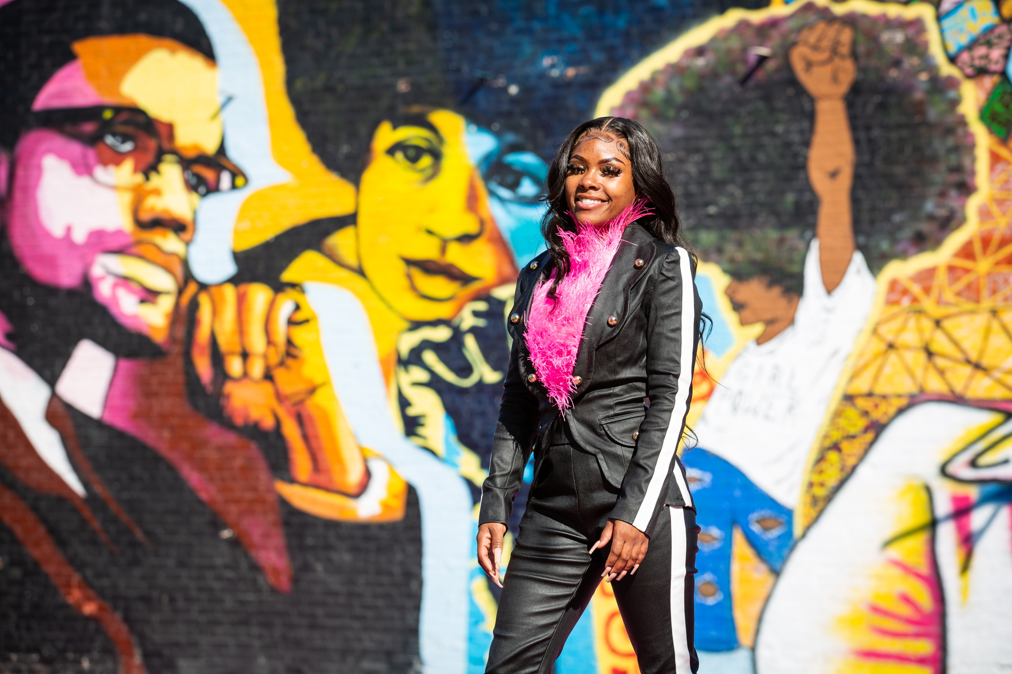 Image: Destine Phillips poses for a portrait at the POP Court public plaza in Chicago's Austin neighborhood. Destine wears black pants and jacket and a pink scarf, and she is looking at the camera while smiling. Behind her is a mural featuring images of Malcolm X, Angela Davis, and a girl with a “girl power” t-shirt raising her left arm and fist into the air. The mural is painted in bright hues of pink, red, orange, yellow, blue, green, and black. Photograph by Kristie Kahns.