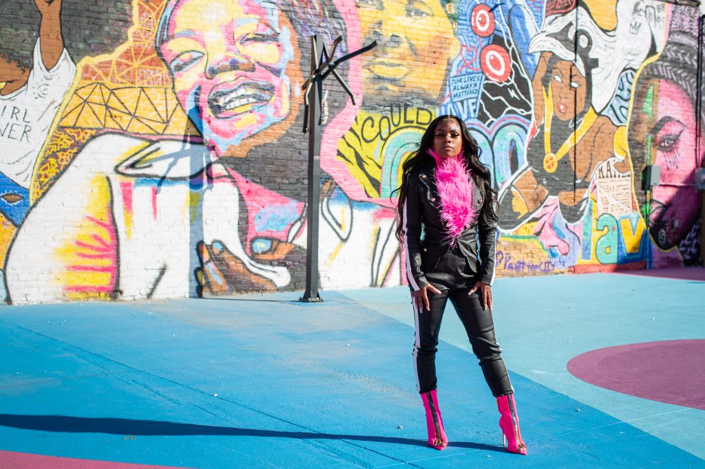 Image: Destine Phillips poses for a portrait at the POP Court public plaza in Chicago's Austin neighborhood. Destine wears black pants and jacket and a pink scarf, and is looking at the camera. In the background, the mural features images of Maya Angelou, the superhero Storm, and a variety of other figures and patterns painted in an array of bright colors. The plaza itself is also painted in shades of blue and purple. POP Court is a site where Phillips and the R.E.A.L. Youth Initiative have previously held some of their community events. Photograph by Kristie Kahns.