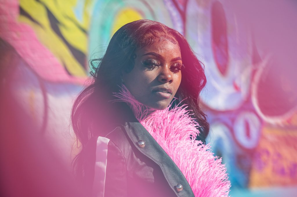 Image: Destine Phillips poses for a portrait at the POP Court public plaza in Chicago's Austin neighborhood. Destine wears a black jacket and a pink scarf, and is looking at the camera. Blurred in the background is a large mural painted in various shades of blue, purple, yellow and green. Photograph by Kristie Kahns.