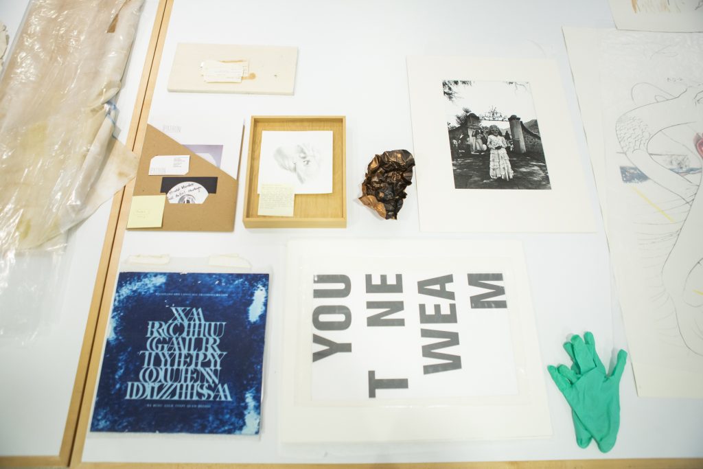 Image: A selection of materials from DePaul Art Museum's Latinx collection. Materials include prints, text-based work, a cd, and works on paper. A pair of mint-colored gloves lay on the table to the bottom right. Photo by Kristie Kahns.