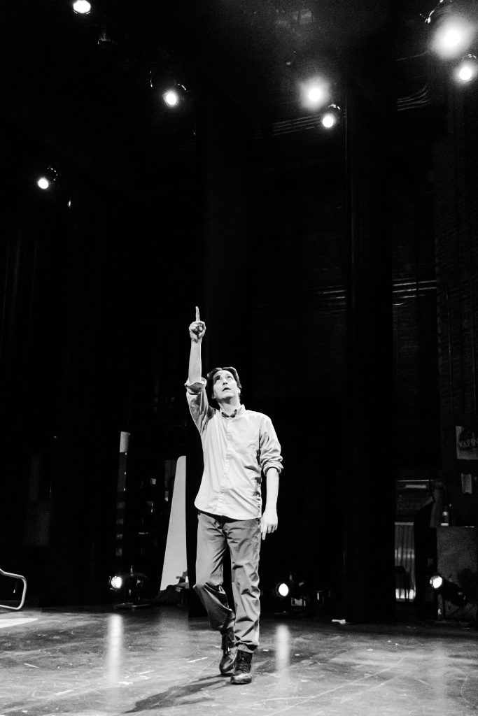 Image: Greyscale photo of Kurt Chiang standing in a theater, pointing his right arm up towards the ceiling lights. Photo by Ella Kang.