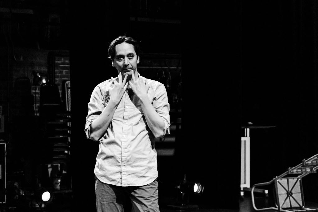 Image: Greyscale image of Kurt Chiang from the legs up in a theater. His hands are over his clavicles, with some fingers touching his chin and cheek. Image by Ella Kang.