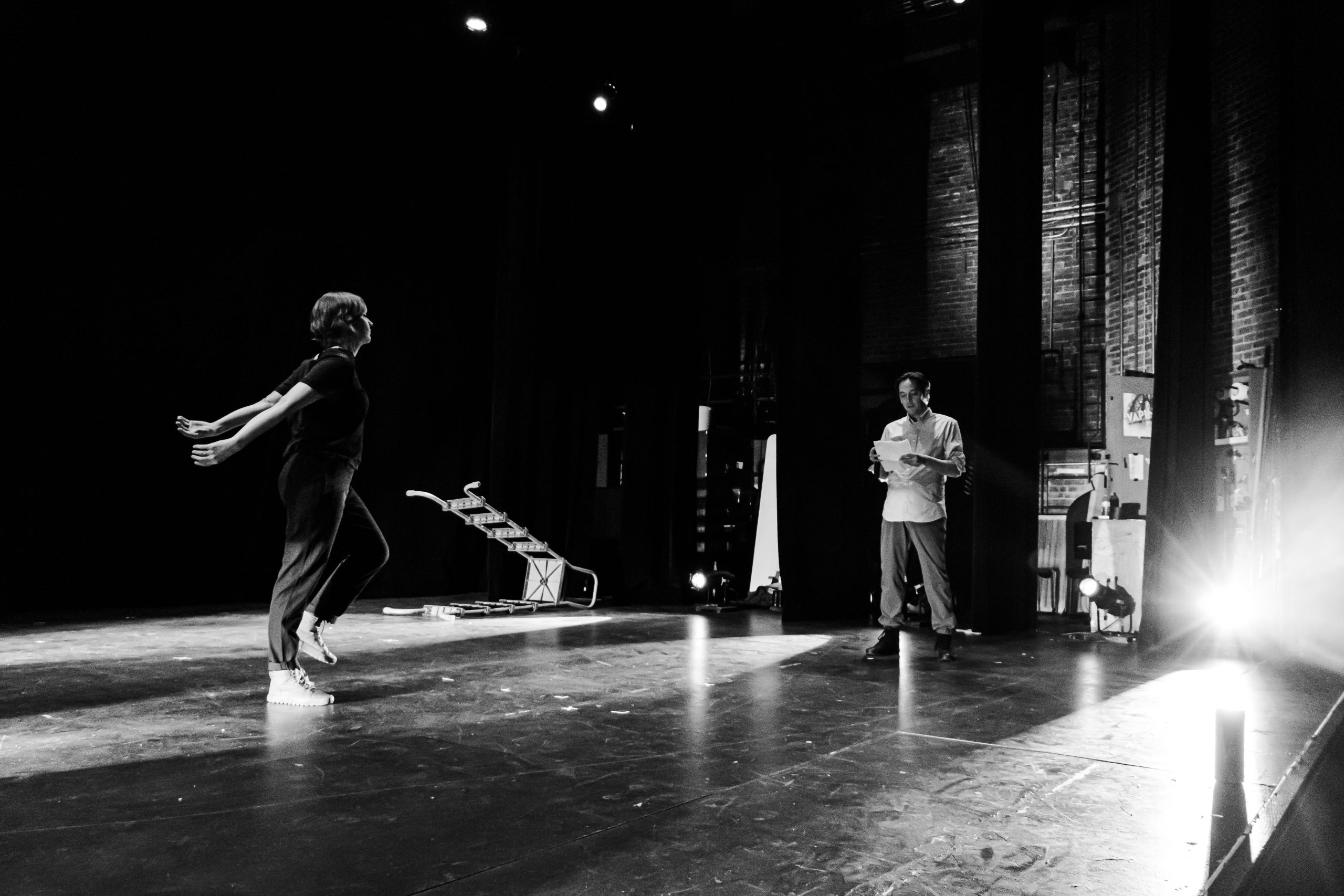 Image: A greyscale photo of Melinda Jean Myers facing Kurt Chiang in a theater. Melinda's arms are outstretched and Kurt is looking at a paper in his hands. There's a bright light behind Kurt and a ladder lying on the floor. Photo by Ella Kang.