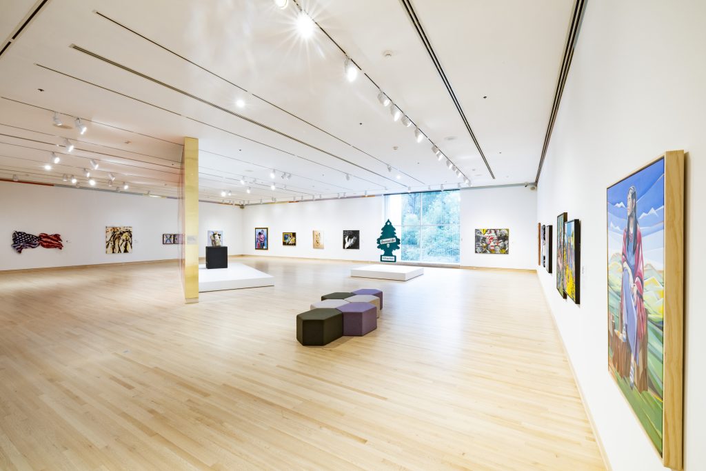Image: Installation view of We. The Culture: Works by The Eighteen Art Collective in the June M. McCormack Forefront Galleries, September 23, 2022–September 24, 2023. Gazing toward the window in a white-walled exhibition space with two platforms for sculpture work and hexagonal seats in the middle. Photo courtesy of the Indianapolis Museum of Art at Newfields.