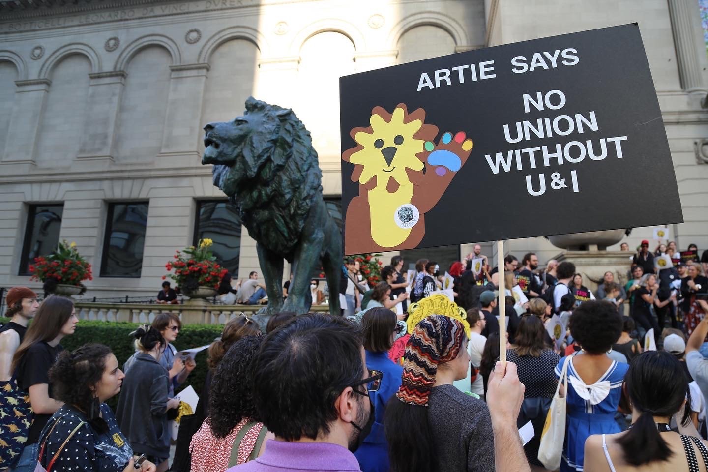 Image: a group of people congregating in front of the Art Institute of Chicago, a grey building. In the mid-ground is an oxidized copper lion sculpture. In the foreground is a person holding a black sign with a cartoon lion that says in white text, "ARTIE SAYS NO UNION WITHOUT US!" Image courtesy of AICWU/AFSCME.