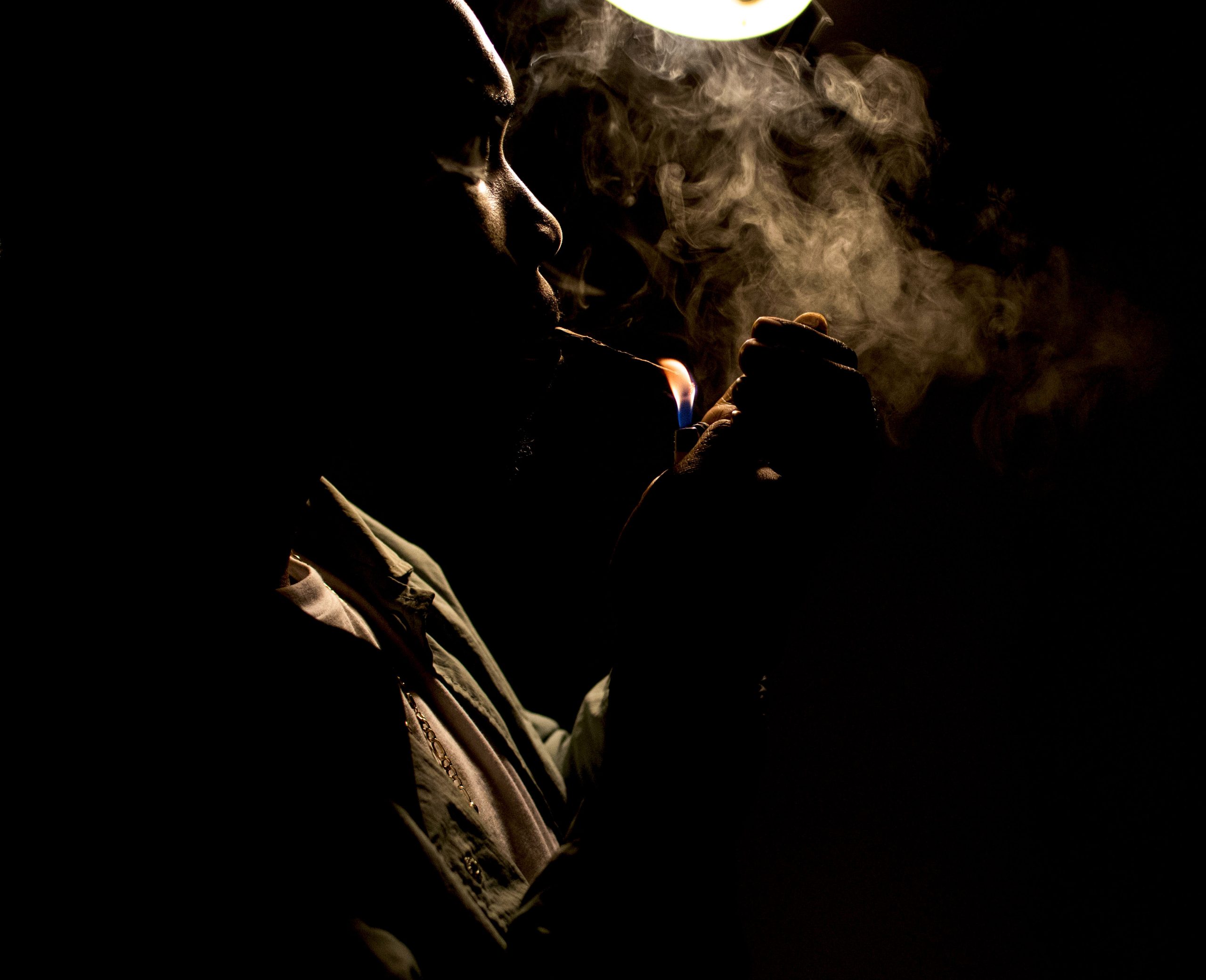 Image: A dark space with a single white lamp over head. In silhouette and smoke, a person cradles a lit match close to the cigarette in their mouth. Photograph by EdVetté Jones.