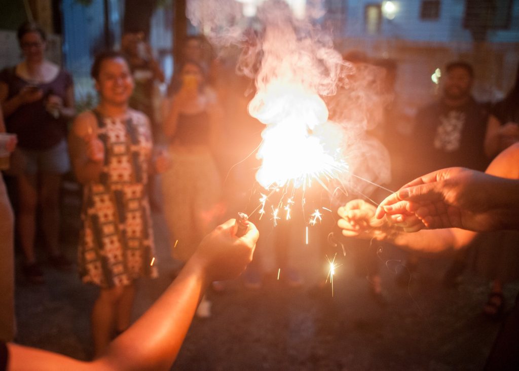 Image: A bright spark can be seen at the center of the image with hands reaching in to light individual sparklers from the flame. In the background different people can be seen smiling. 