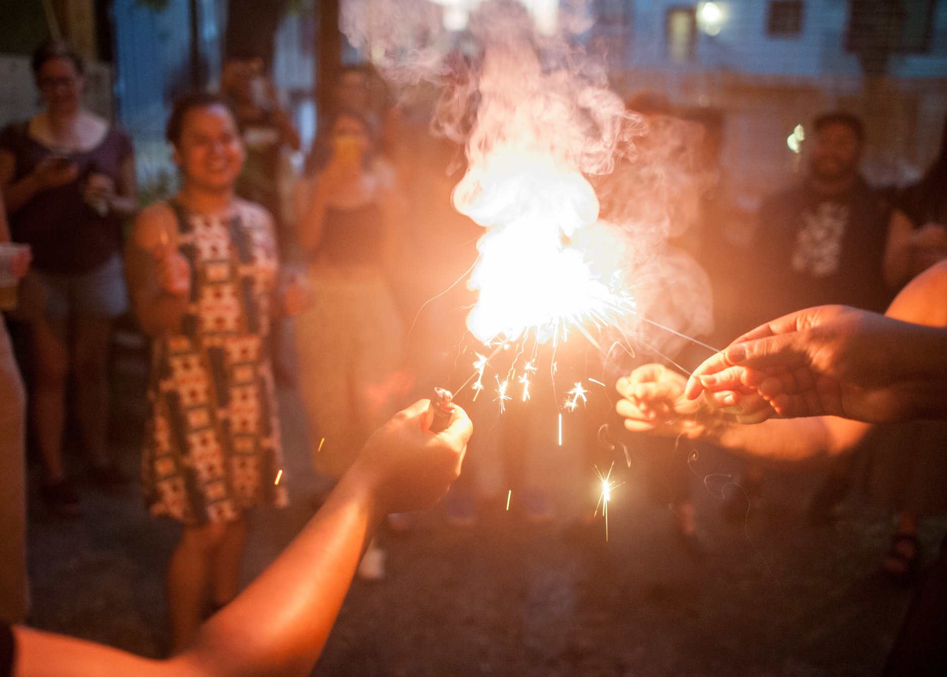 Image: A bright spark can be seen at the center of the image with hands reaching in to light individual sparklers from the flame. In the background different people can be seen smiling.