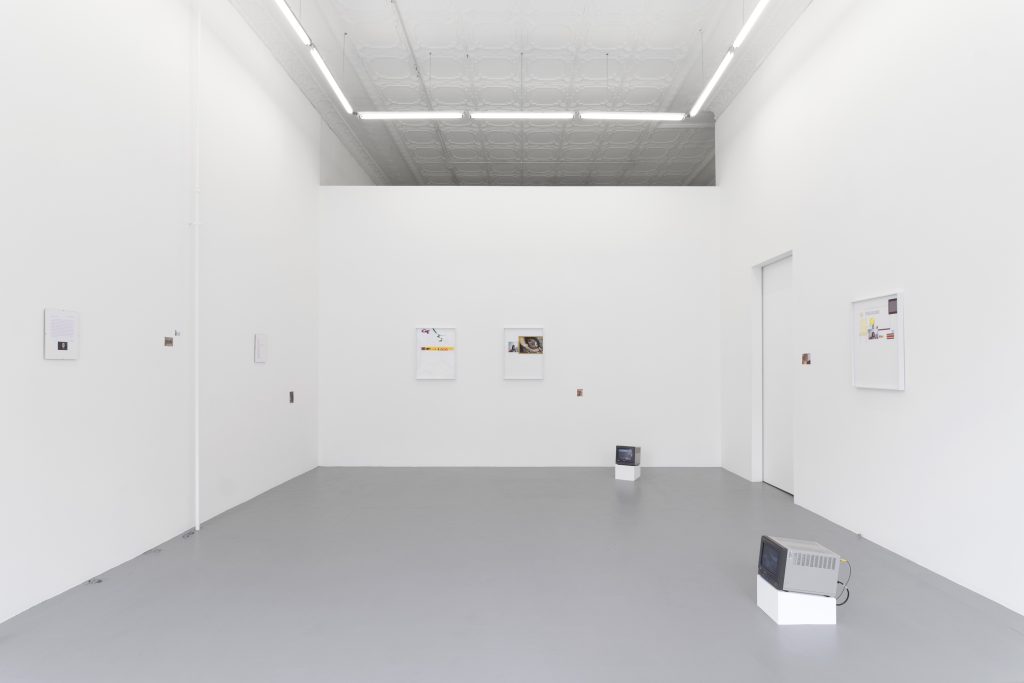 A white-walled gallery space with several framed works hung on each wall and two CRT monitors on the right side of the room.
