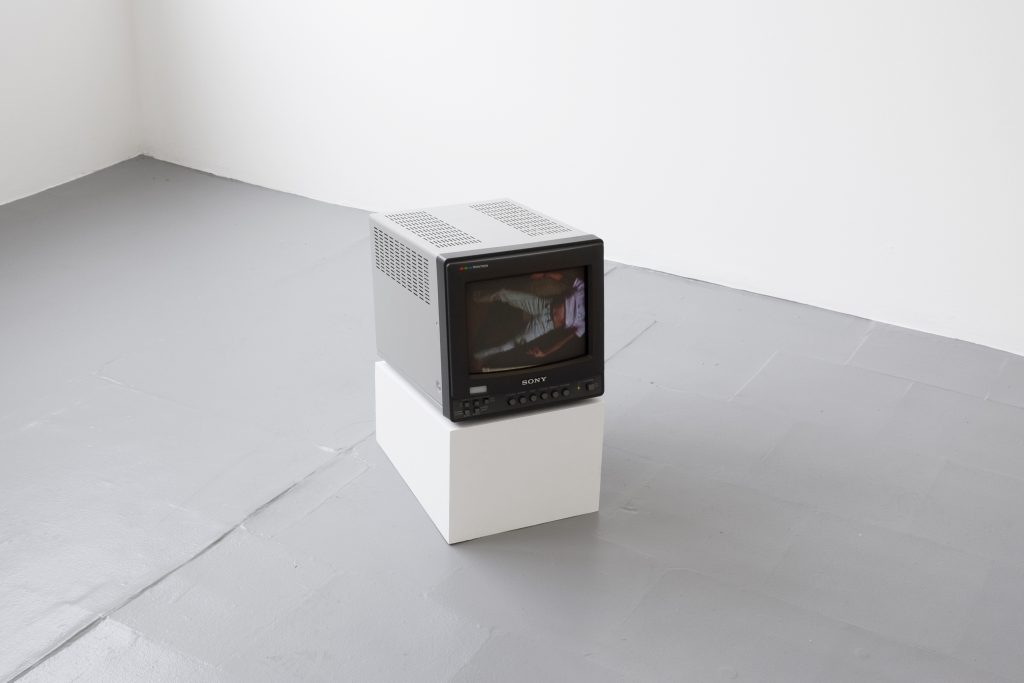 In a corner of the gallery, a Sony CRT monitor plays a video of a figure wearing jeans, a purple shirt rolled up to reveal their stomach. The figure's head is out of frame.