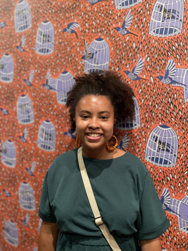 Image: A photo of the author smiling and facing towards the camera. She is in front of a Bisa Butler portrait quilt depicting purple-colored birds flying out of or trapped inside cages on a pink-salmon colored background of grass at the Art Institute of Chicago. Image courtesy of Chenoa Baker.