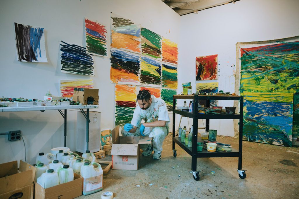 Image: Paul Verdell is bent over a box in his studio. He is surrounded by various art supplies, and behind him on the wall are various vibrant, abstract works of his. Photo by Kai Newby. Image courtesy of the artist and Library Street Collective. 