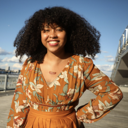 Image: A headshot of Chenoa Baker, who smiles at the viewer with a hand on her hip.