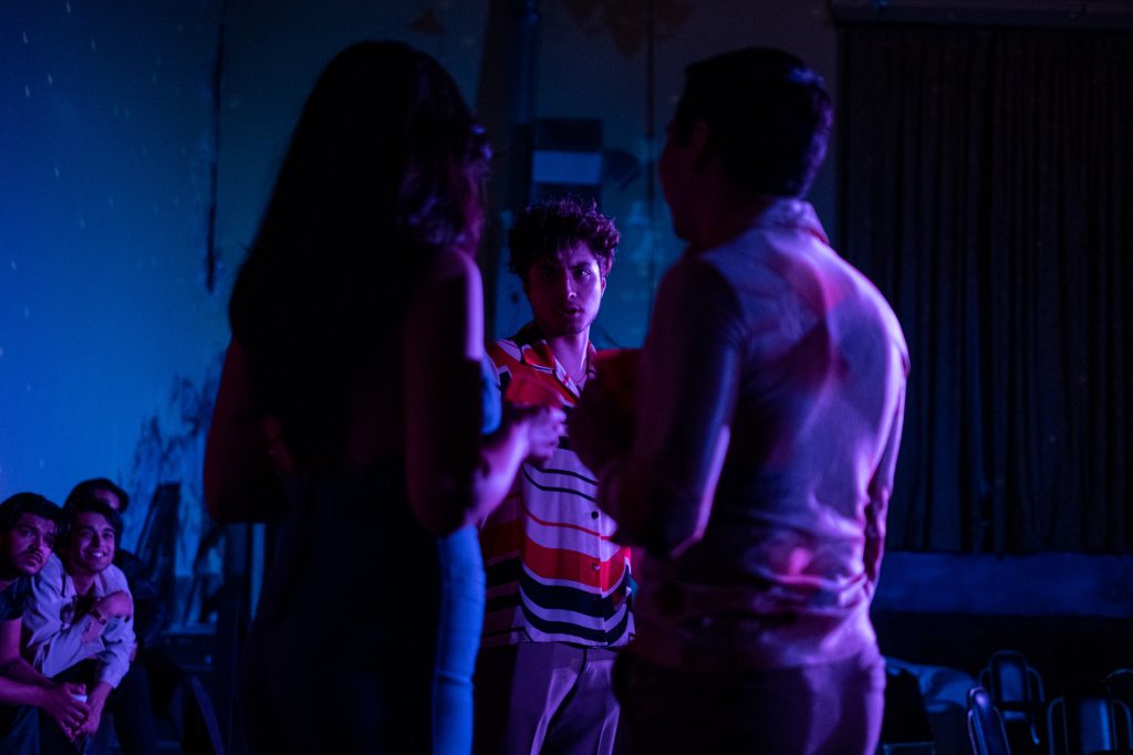 Image: Three people stand on stage during the performance of 'The Wizards.' One person is looking straight ahead while the other two have their backs to the viewer. The scene is darkly lit with blue lighting. Photo Courtesy of Chicago Latino Theater Alliance.