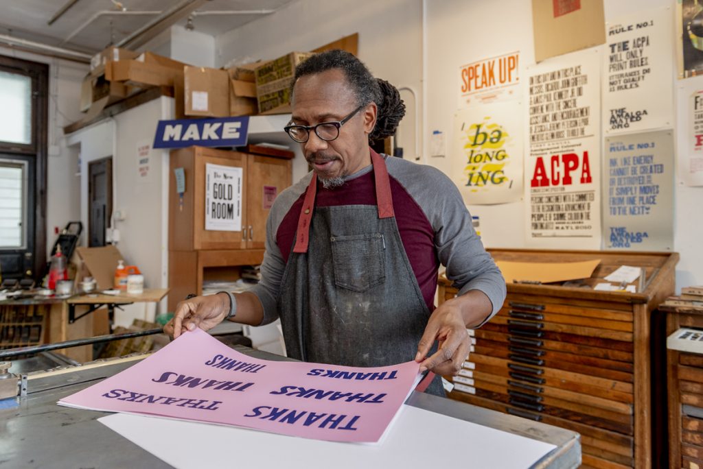 Image: Artist Ben Blount standing over a table in his studio, holding a letterpress print that says "thanks" in six different typefaces. In the background you can see posters and prints hanging on the wall above wooden shelves and flat files. Photo by Ryan Edmund Thiel.