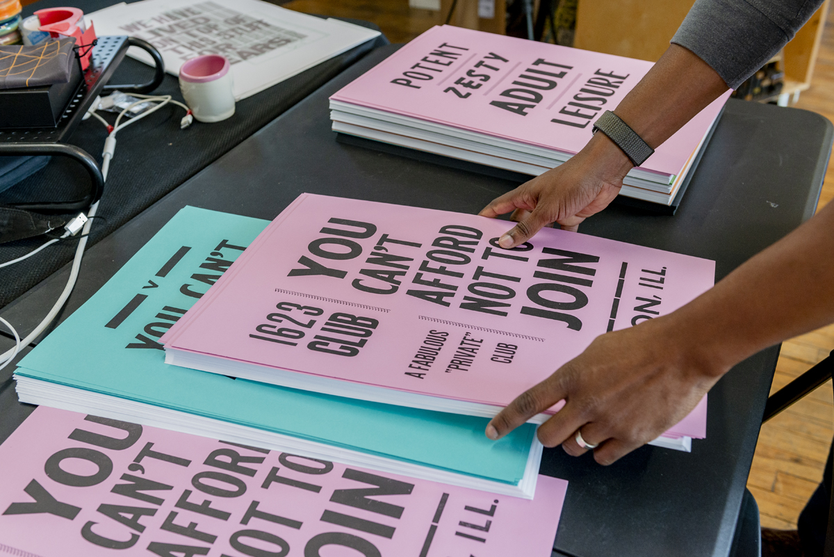 Image: A print on pink paper can be seen with the words "You Can't Afford Not To Join" in bold type. Around it are other prints, some that are also on pink paper, others that are printed on teal. Artist Ben Blount's hands enter into the frame from the right, holding onto a stack of prints. Photo by Ryan Edmund Thiel.