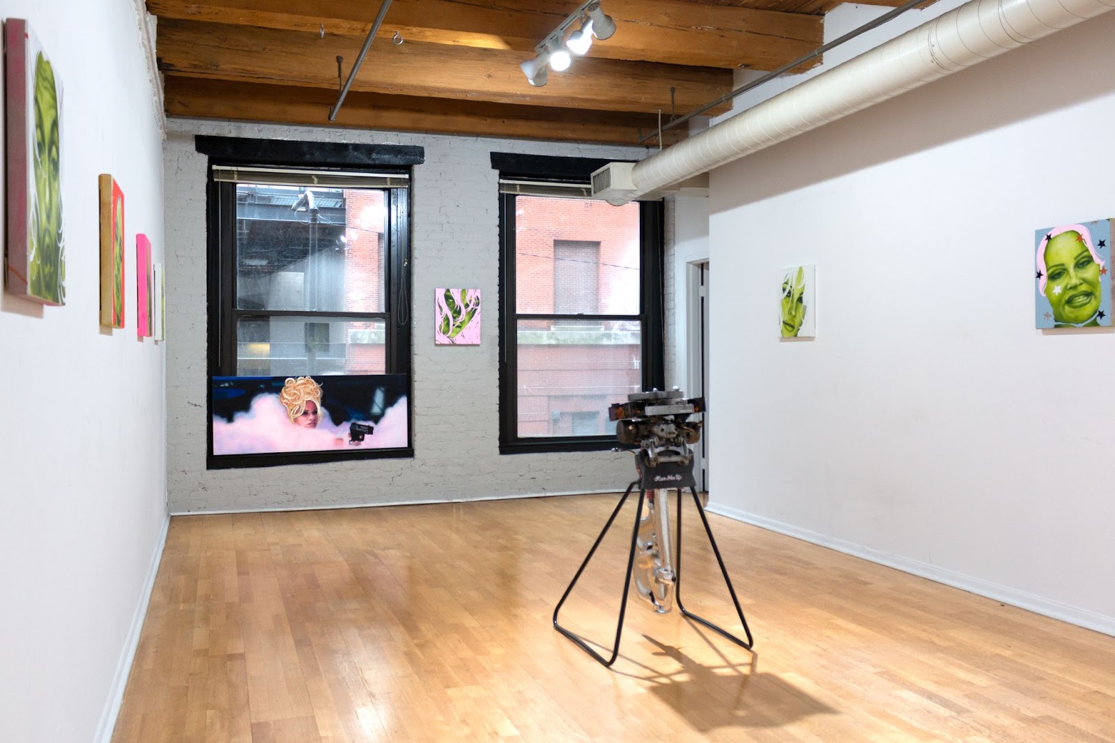 Image: Exhibition view of Liza Jo Eilers' The Care and Keeping of You. In the center of the exhibition space stands a boat motor. Airbrushed portraits of actresses Jennifer Coolidge and Pamela Anderson are hung on the walls surrounding the motor. On the right of the image, a painting of Coolidge rendered in sickly chartreuse green dominates the white wall. To the back of the image, near the windows, Pamela Anderson emerges in frame within a pink cloud of smoke while holding a gun. Courtesy of artist. 
