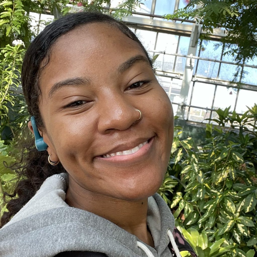 Image: A photo of Tianna Garland, who smiles at the camera with plants behind her.