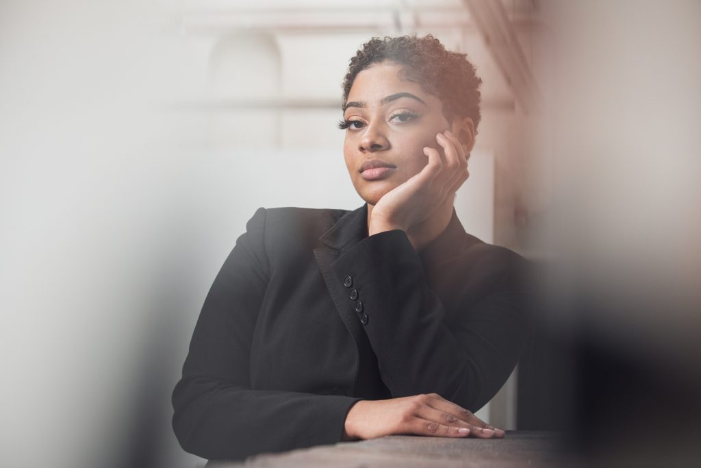 Image: A portrait of Saidayah Kirk standing, leaning on a surface, looking into the camera. Her hand rests at her chin and her other hand rests on the surface she's leaning on. She is framed by blurry edges in the foreground. Photo by Kristie Kahns.