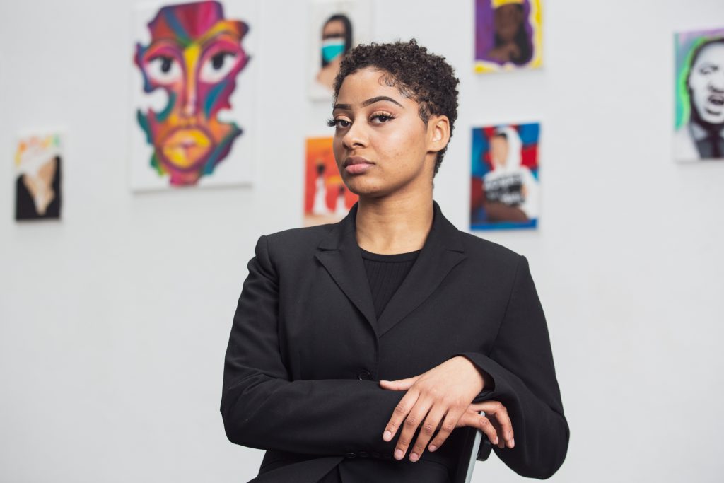 Image: Saidayah Kirk sits on a chair in front of a wall displaying several of her paintings, drawings, and mixed media works. She is dressed in all black and her hands are crossed at the wrist and resting on the back of a chair. Photo by Kristie Kahns.