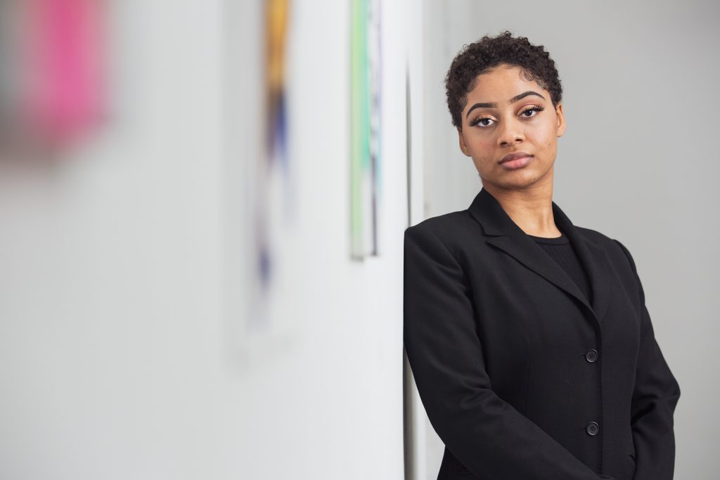 Image: A portrait of Saidayah Kirk standing, leaning against a wall, looking into the camera. Blurred out on the wall next to her are small glimpses of some of her paintings. Photo by Kristie Kahns.