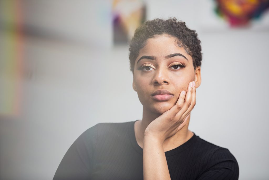 Image: A portrait of Saidayah Kirk from the shoulders up. She is looking directly into the camera and her hand rests at her chin. Behind her you can see blurry images of her artworks hanging on the wall. Photo by Kristie Kahns.