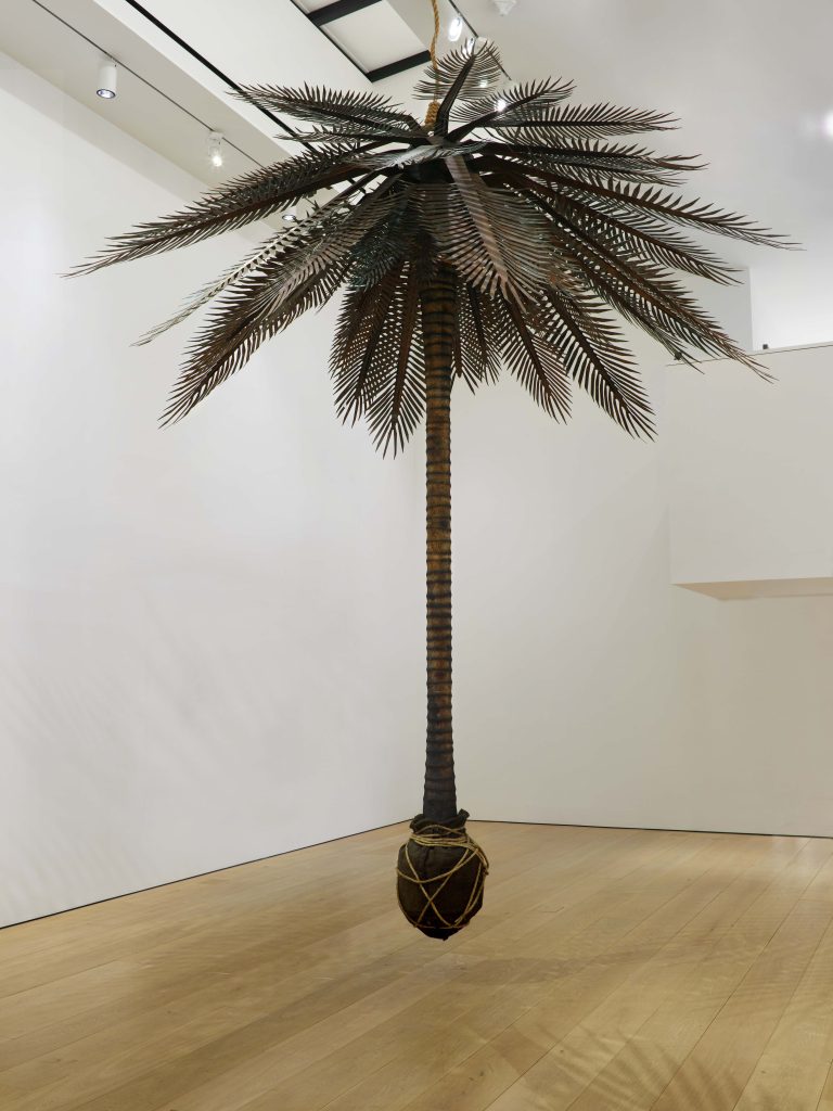 Image: Teresita Fernández, Rising (Lynched Land), 2020. Copper, wood, burlap, rope. 192 × 176 × 176 in. (487.7 × 447 × 447 cm). A palm tree is suspended from the ceiling over a wooden floor and in front of white walls. Courtesy of the artist and Lehmann Maupin. 