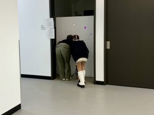 Image: Two people, bent over peering into a white plaster wall of holes at the exhibition "Glory Hole". Image courtesy of David Downs.