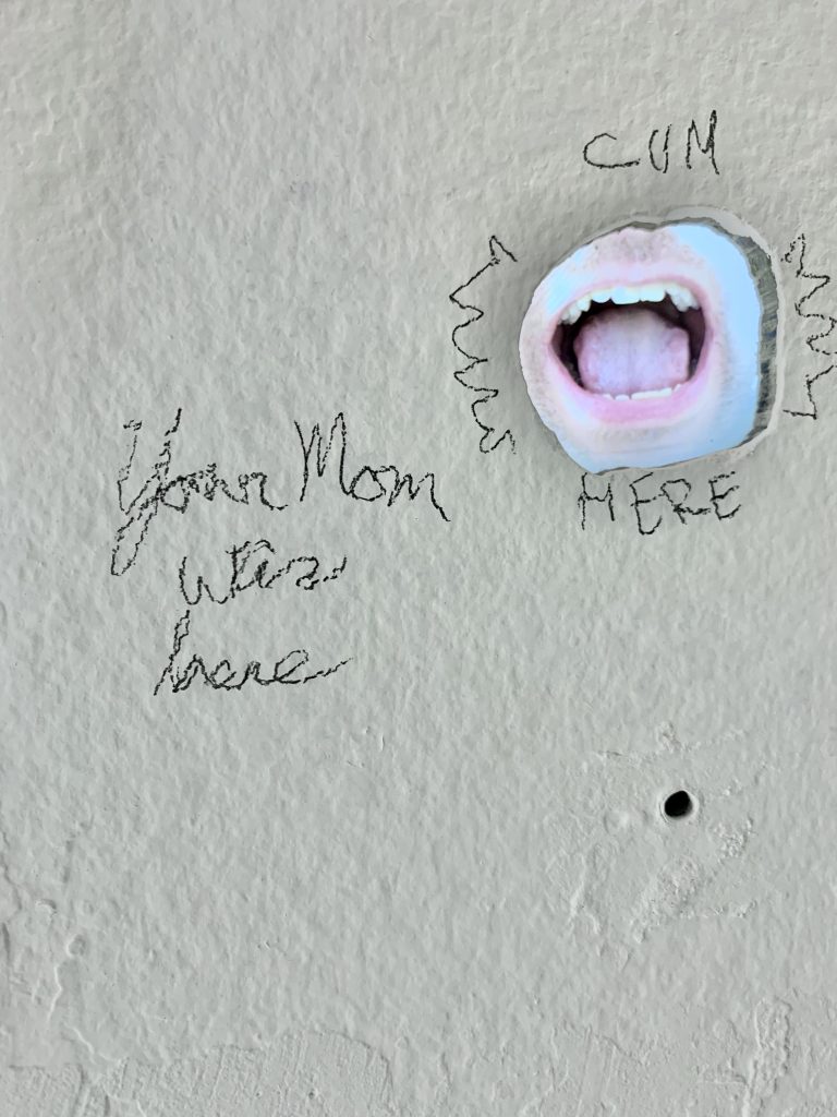 Image: David Downs, Selfie Hole, 2022. A hole in a white plaster wall. Written above the hole is the word "CUM" and written below the hole is the word "HERE". Visible through the hole is an open mouth. Written next to the hole are the words "Your mom was here". Images provided by David Downs.