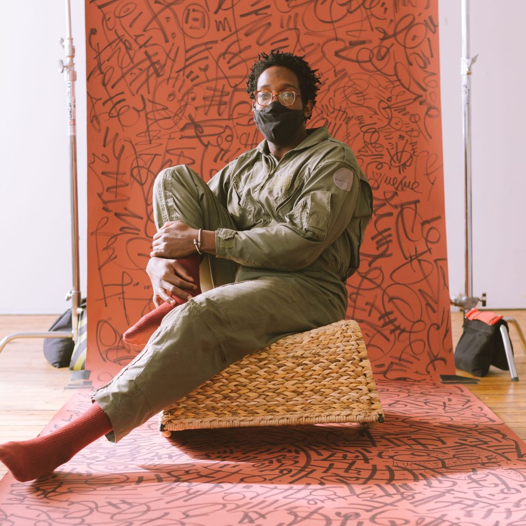 Image: A fully body photo of a person wearing an olive jumpsuit, black mask, glasses, and clay colored socks. They sit on a woven chair against a clay colored wall with black scribbling on it.