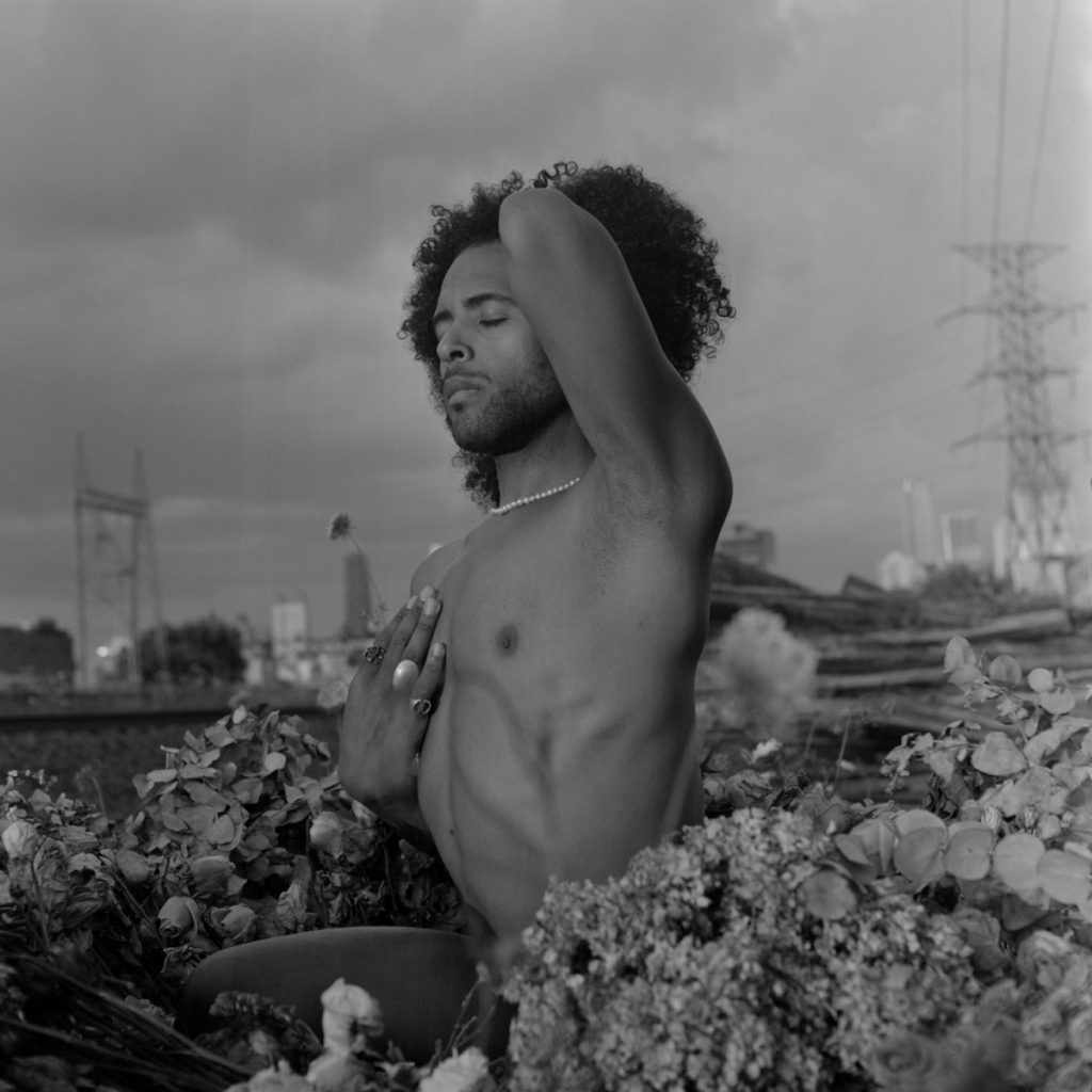 Image: A black-and-white portrait of Erol turned to the left, eyes closed, right hand at his heart, and left arm raised. He is sitting among dry flowers outdoors on an elevated train track. Buildings, electricity towers, and a cloudy sky can be seen in the background. Photo by Ryan Edmund Thiel.