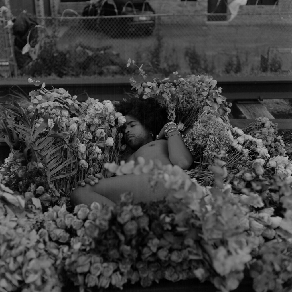 Image: A black-and-white portrait of Erol reclined on the train tracks among dry flowers, outdoors on an elevated train track. A chain-link fence can be seen in the background. Photo by Ryan Edmund Thiel.