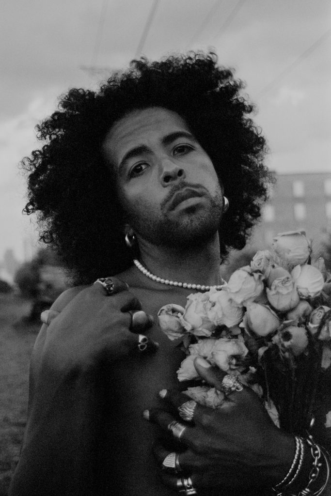 Image: A close black-and-white portrait of Erol standing with one hand at his shoulder, wearing a string of white pearls, and holding a bouquet of dry flowers, outdoors. Buildings can be seen in the background. Photo by Ryan Edmund Thiel. 