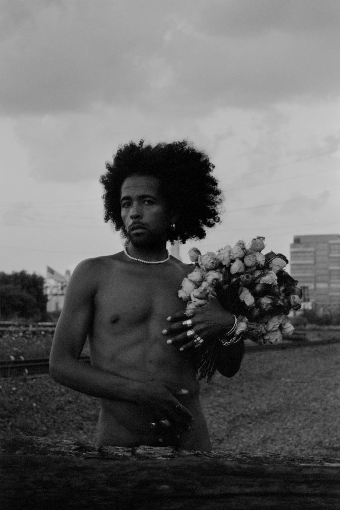 Image: A black-and-white portrait of Erol seen from the waist up, standing with one hand at his stomach, wearing a string of white pearls, and holding a bouquet of dry flowers, outdoors. A wooden railway sleeper can be seen in the foreground and buildings can be seen in the background. Photo by Ryan Edmund Thiel.