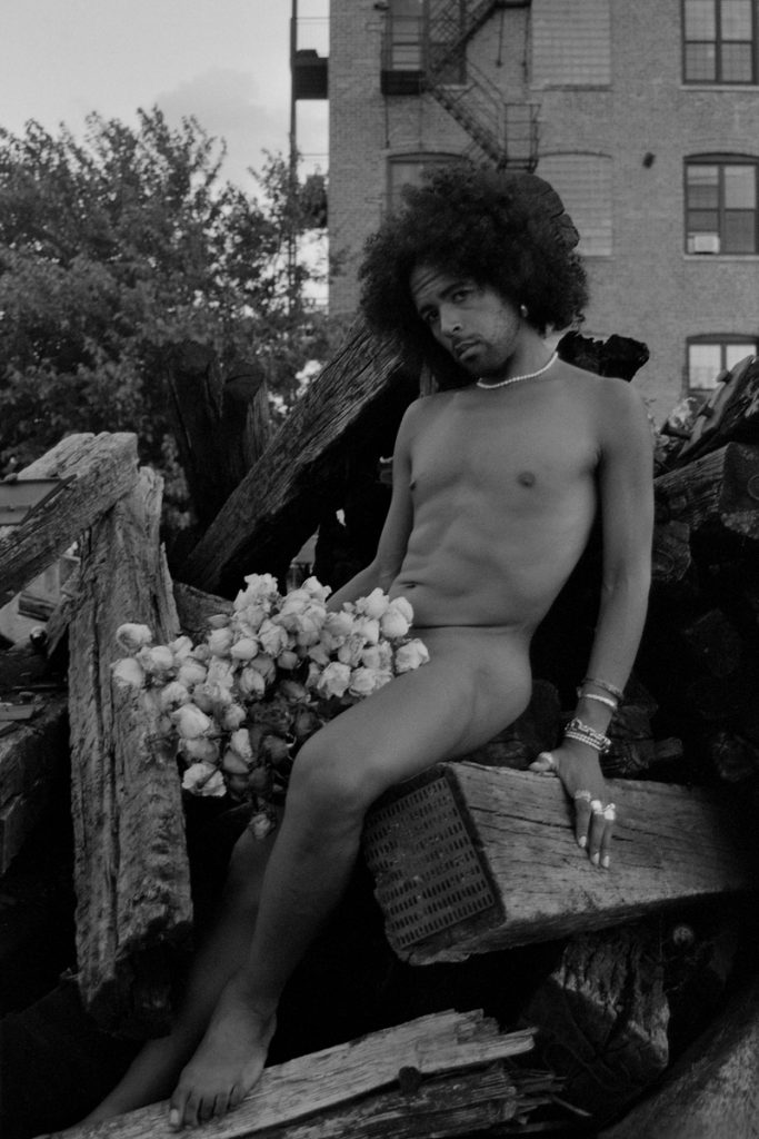Image: A black-and-white full-body portrait of Erol sitting on a series of wooden railway sleepers with a bouquet of dry flowers on his lap. Trees and buildings can be seen in the background. Photo by Ryan Edmund Thiel.